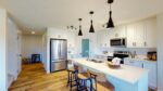 A kitchen in a home with white, shaker style cabinets, chevron patterned white tile backsplash, white counter tops, black pendant lights over the island and warm wood floors.