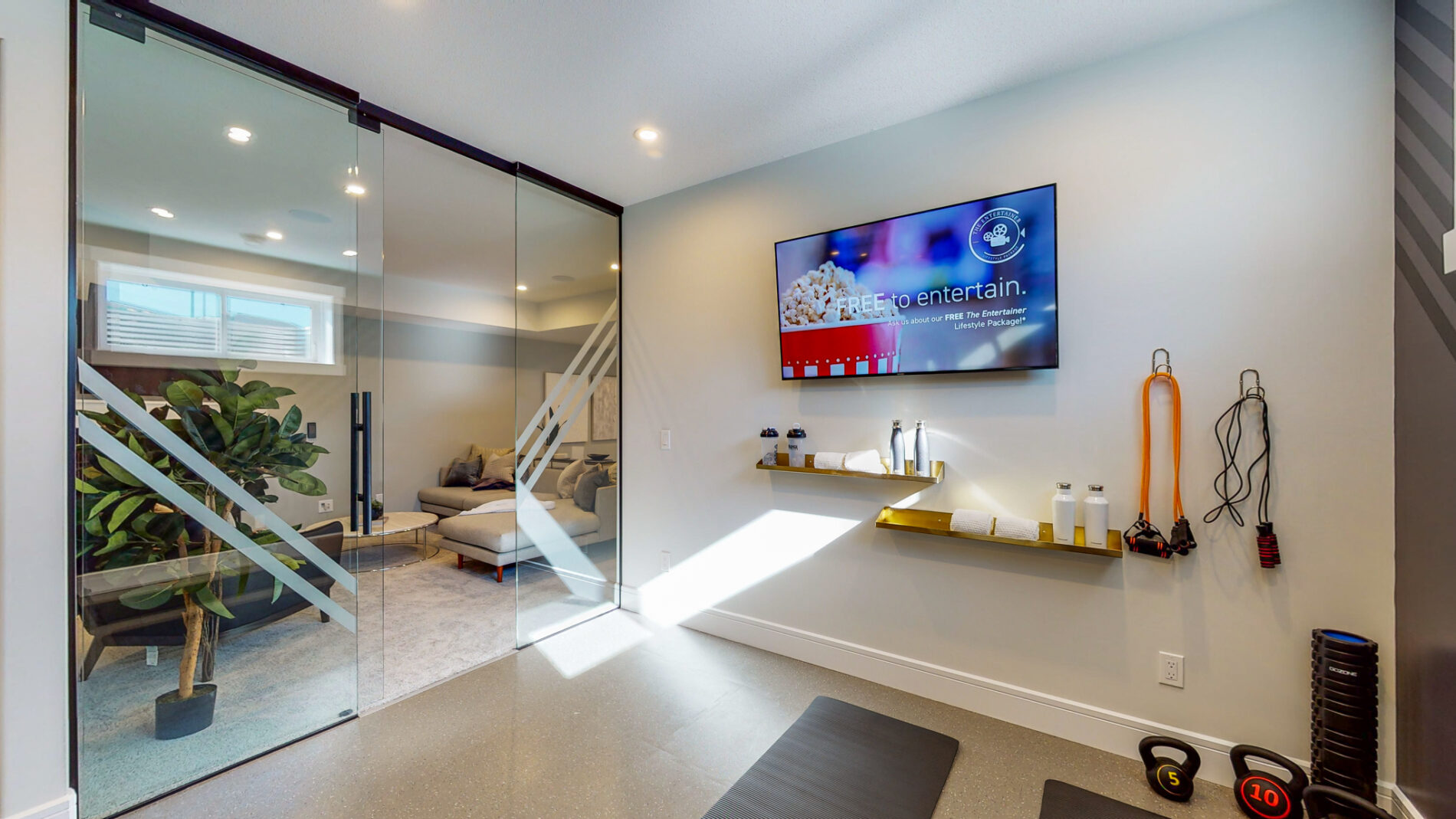 Home gym with glass wall and door separating it from the family room, rubber flooring, and tv and shelves on the wall