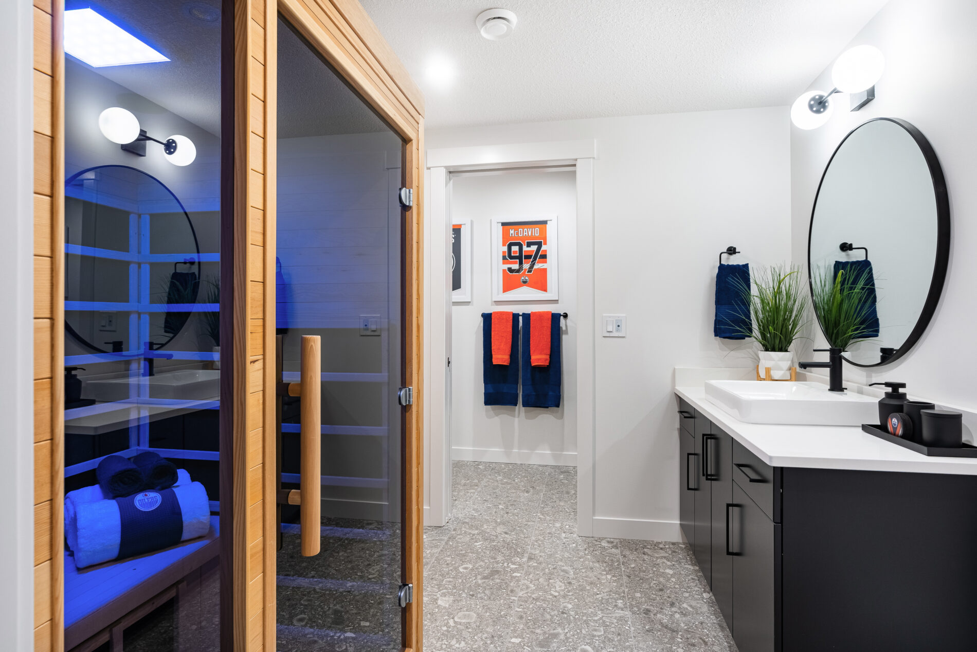 Bathroom of Oilers Fan Cave basement with large cedar sauna to the left, a sleek black vanity with round mirror above and looking through to the shower room with blue and orange towels hanging