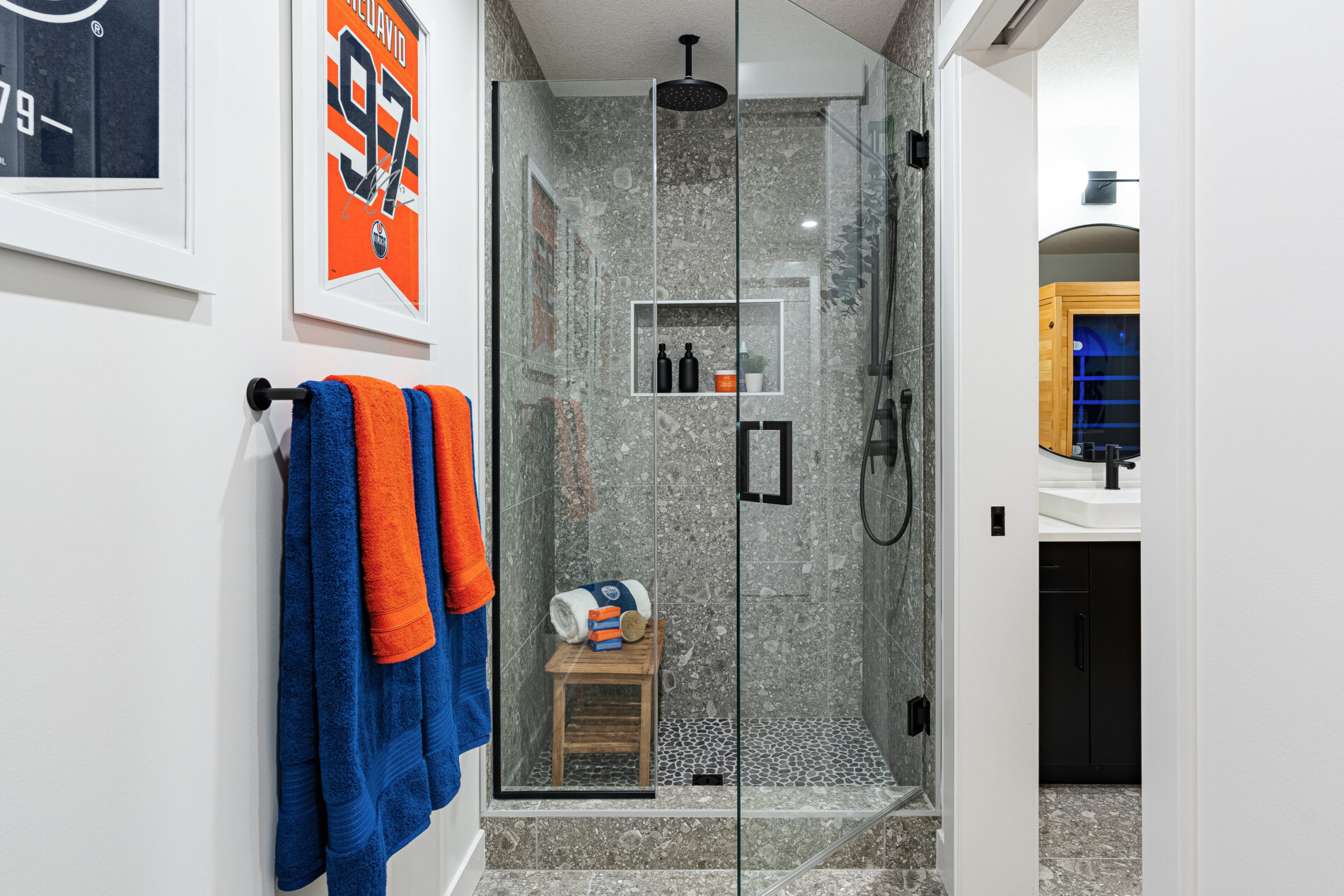 Spa like shower in Oilers Fan Cave basement shower room with grey tile to the ceiling, glass enclosure, black fixtures with blue and orange towels and accents