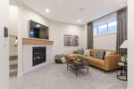 A fireplace with white tile with a warm wood mantle is the focal point of the photo. A cozy couch is placed to the side of the fireplace in front of the window in the basement of the home.